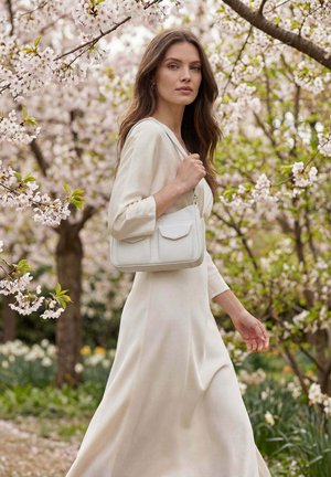 Femme en longue robe crème portant un sac à main blanc, marchant dans un jardin avec des cerisiers en fleurs.