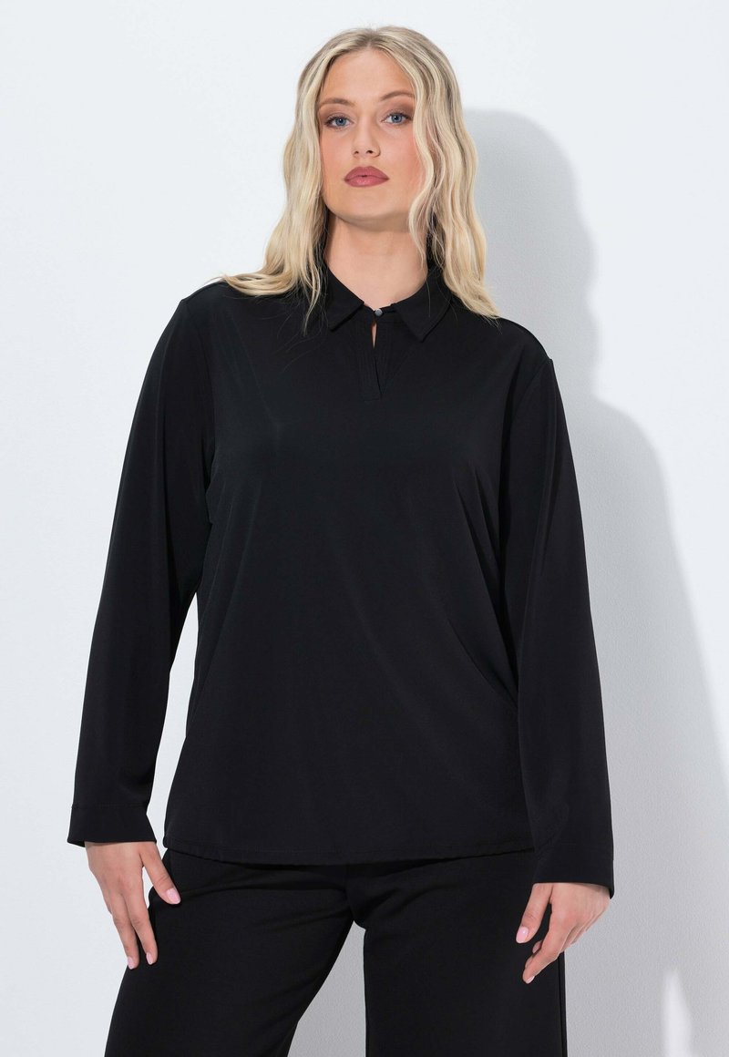 Blusa nera a maniche lunghe con collo a bottoni e un sottile dettaglio plissettato sullo scollo, realizzata in tessuto morbido. Abbinata a pantaloni neri.