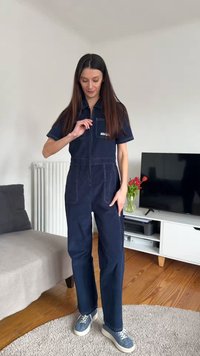 Kvinde i mørkeblå overall og mønstrede sneakers står i en stue med et TV, et hvidt skab, en grå sofa og blomster på skabet.