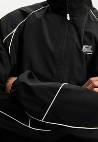Zwarte, lichte jas met een hoge kraag, een ritssluiting aan de voorkant en witte accentpaspels langs de naden. Voorzien van een logo op het borstgebied.