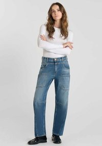 Jeans en denim bleu clair avec une coupe droite, des poches avant et des coutures visibles, assortis à un haut blanc à manches longues et des chaussures noires.