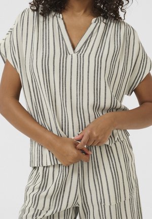 Woman adjusting a beige and black vertically striped short-sleeve top, matching striped pants visible from waist down.