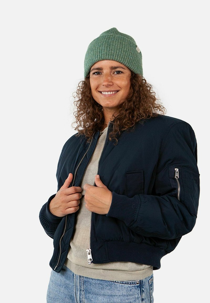 Navy bomber jacket with ribbed cuffs and hem, featuring zippered and patch pockets. Worn with a green ribbed beanie and gray shirt underneath.
