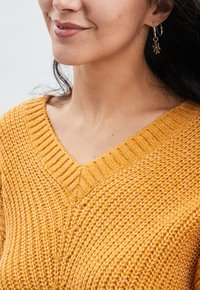 Senfgelber Strickpullover mit tiefem V-Ausschnitt, der ein strukturiertes Muster aufweist. Silberne Ohrhänger mit einem Blatt-Anhänger sind sichtbar.