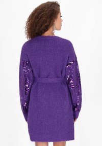 Fioletowy kardigan z teksturowanym ciałem i rękawami pokrytymi cekinami. Posiada pasek w talii i luźny krój, idealny do noszenia na warstwy.