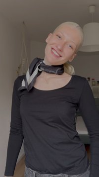 Sort langærmet skjorte i blødt stof; sort og hvidt mønstret tørklæde bundet omkring halsen; lys hud og lyst blondt hår.