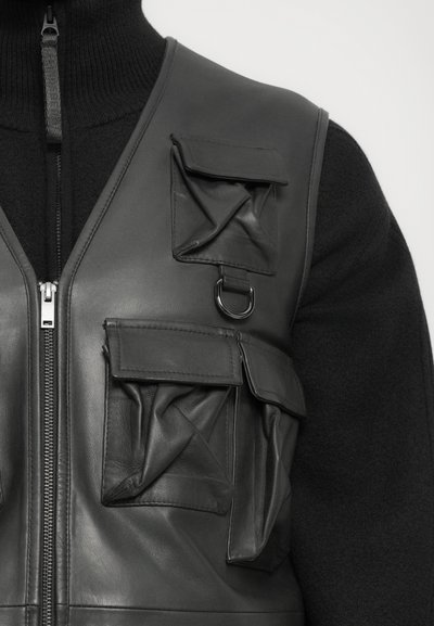 Close-up of black leather vest with multiple flap pockets and silver ring over black zip-up sweater.