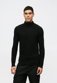 Juodas megztinis su aukštu kaklu, ilgomis rankovėmis, pagamintas iš minkštos, tekstūrinės medžiagos. Turi pusiau užtrauktuką ant apykaklės, prilipnanti siluetas.