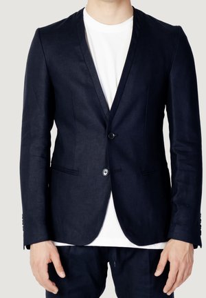 Mann trägt einen dunkelblauen Blazer mit zwei Knöpfen über einem schlichten weißen T-Shirt mit Rundhalsausschnitt, Hände entspannt an den Seiten.