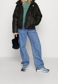Sort pufjakke, grøn sweater og løse blå jeans. Iført blå og grøn sneakers og holder en sort faux fur taske.
