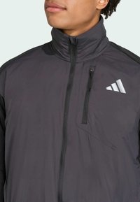 Black lightweight jacket with a high collar, zippered pocket, and reflective logo. Material appears smooth and water-resistant.