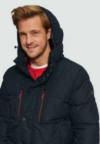 Glunderende man in een zwarte gewatteerde jas met capuchon over een rode trui, recht vooruit kijkend tegen een effen lichte achtergrond.