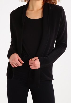 Femme portant un blazer noir texturé ouvert sur un haut noir et un pantalon noir, tenant les bords du blazer avec les deux mains.