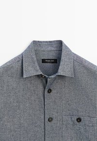 Grijs gemêleerd overhemd met knopen, voorzien van een klassieke kraag, borstzak en zwarte knopen tegen een gestructureerde stofachtergrond.