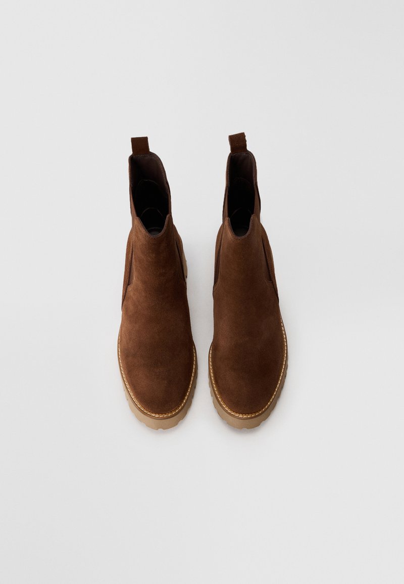 Brązowe zamszowe botki Chelsea z beżowymi gumowymi podeszwami, elastycznymi panelami bocznymi i pętlami do zakupu, pokazane z góry na białym tle.