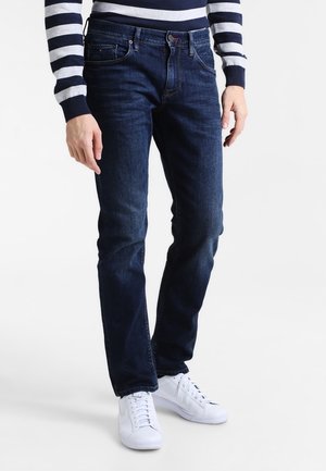 Jeans de mezclilla azul oscuro con corte recto, cinco bolsillos y una textura sutil. Llevados con zapatillas blancas y una camiseta de rayas en azul marino y gris.