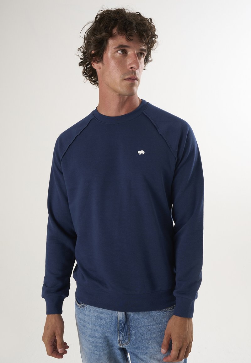 Mörkblå sweatshirt i mjukt bomullstyg, med rundad halsringning, raglanärmar och en liten vit björnlogotyp på bröstet.
