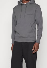 Šedá mikina s klokánkovou kapsou, nastavitelnou kapucí se šňůrkou a decentním logem. Vyrobena z měkkého, hladkého materiálu.