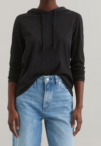 Persona che indossa un hoodie nero a maniche lunghe e jeans chiari a vita alta, con le mani rilassate ai lati su uno sfondo neutro.