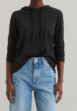 Une personne portant un hoodie noir à manches longues et un jean taille haute bleu clair, les mains détendues sur les côtés devant un fond neutre.