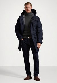 Casaco acolchoado azul-marinho com interior cinzento, usado sobre um suéter escuro e jeans escuros. O modelo está em pé com as mãos nos bolsos, calçando mocassins castanhos.