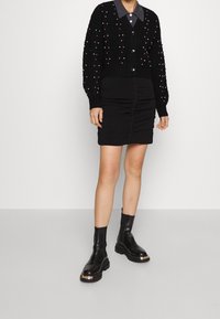 Cardigan noir en tricot avec des accents en perles roses sur une robe noire froncée. Paire de bottines noires à semelles épaisses avec des détails métalliques.