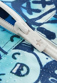 Dragkedjor i tyg i nyanser av blått med ett mönstrat design. Vit plastdragkedja med en stjärnformad dragtab, vattendroppar synliga på ytan.