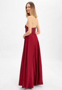 Femme en robe de soirée rouge bustier avec corsage orné, debout légèrement tournée, regardant par-dessus l'épaule sur un fond uni.