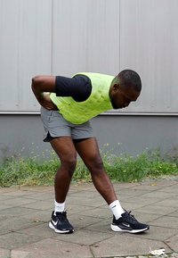 Chaleco acolchado ligero de color verde neón sobre una camiseta negra de manga corta, pantalones cortos grises y zapatillas de running negras, con una persona estirándose hacia adelante.