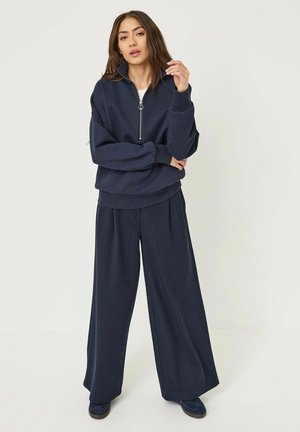 Navy zip-up sweatshirt with dropped shoulders and ribbed cuffs, paired with wide-leg pinstripe trousers in matching navy. Casual footwear completes the look.