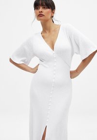 Robe blanche texturée avec un décolleté en V, des manches larges et courtes, et des boutons sur le devant. Présente une fente jusqu'à la cuisse.