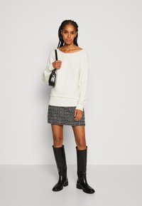 Pull en maille côtelée crème, jupe à motifs noir et blanc, et bottes en cuir noir jusqu'aux genoux. Le mannequin tient un petit sac à main noir.