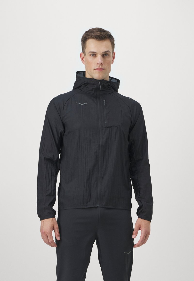 Veste légère noire avec capuche, dotée d'une poche frontale zippée, d'un tissu texturé et d'accents de logo réfléchissants. Manches et ourlet protégés.