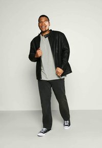 Smiling man standing, wearing a black leather jacket, gray shirt, black pants, and black-and-white sneakers against a plain light background.