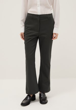 Personne portant un pantalon noir à jambes droites, des mocassins noirs et une chemise blanche à manches longues boutonnée, debout devant un fond uni.