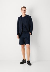 Blazer navy su una maglietta coordinata, abbinato a pantaloni corti navy. Scarpe nere stringate. Tessuto liscio con vestibilità su misura e stile casual.