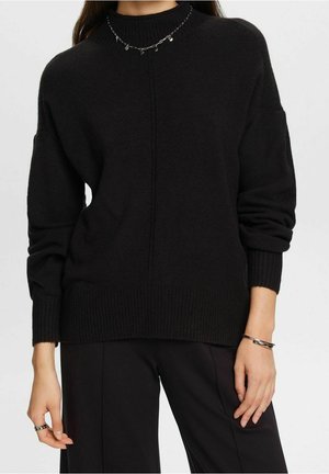 Person trägt einen schwarzen, gerippten Pullover mit langen Ärmeln, kombiniert mit schwarzen Hosen und silbernem Schmuck am Hals, Handgelenk und an den Fingern.