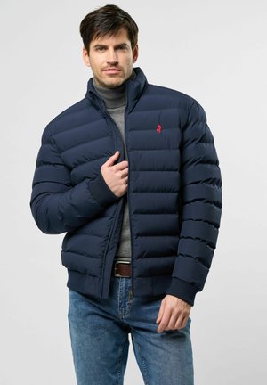 Man in een marineblauwe pufferjack over een grijze coltrui en blauwe jeans, terwijl hij de rits van zijn jack aanpast tegen een effen lichte achtergrond.