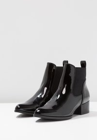 Bottines noires à finition vernie brillante, avec des panneaux latéraux élastiques et un petit talon carré. Elles présentent une texture lisse et une pointe arrondie.