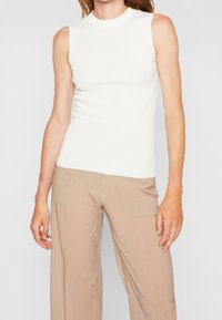 Mujer vestida con una camiseta sin mangas de punto blanco y pantalones beige de corte holgado, de pie contra un fondo gris claro liso.