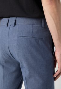 Personne portant un pantalon habillé bleu ajusté et une chemise noire, montrant la poche arrière et les passants de ceinture.