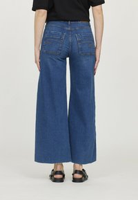 Jean en denim à jambe large de couleur bleu foncé, avec une taille haute, deux poches arrière avec des détails cousus, et des bords effilochés à l'ourlet.