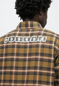 Καρό πουκάμισο από φανέλα, κυρίως κίτρινο μουστάρδας με ναυτικό μπλε και πορτοκαλί στοιχεία. Φέρει τη λέξη «JORDAN» με έντονα, φθαρμένα λευκά γράμματα στην πλάτη.