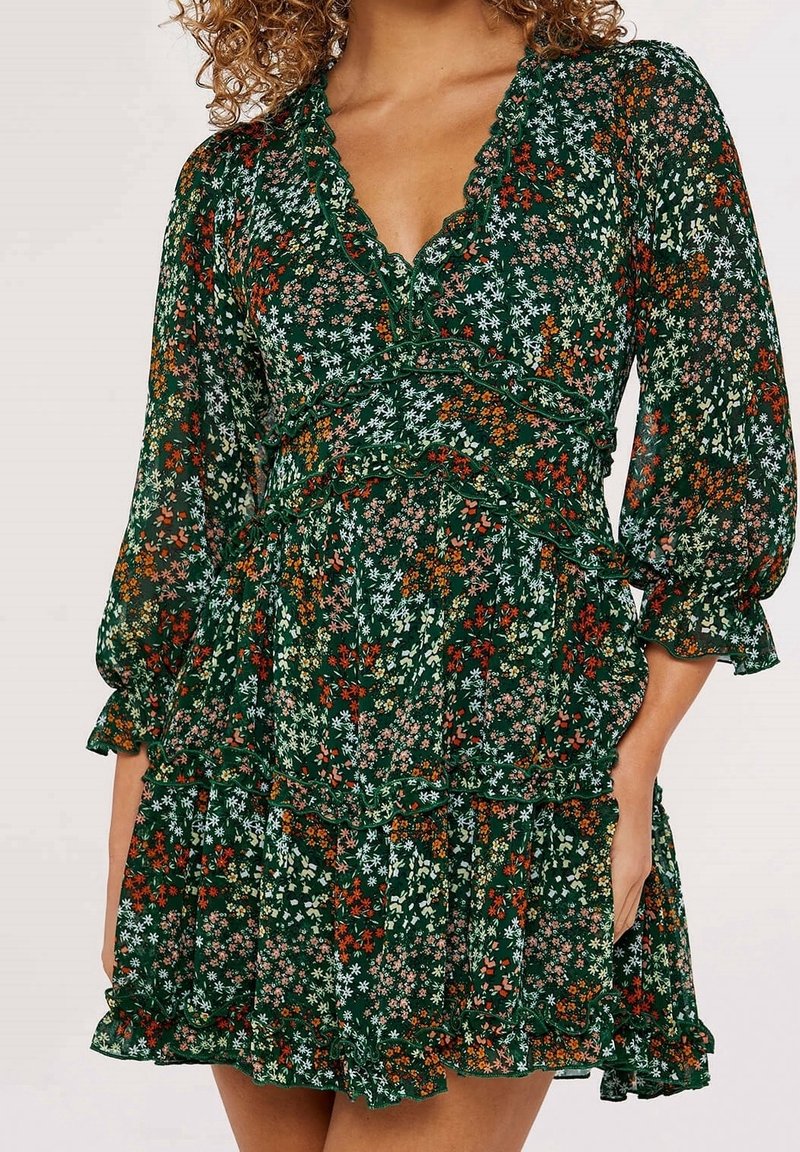 Robe florale verte avec des manches à volants et une jupe à volants, ornée de petites fleurs blanches, orange et roses sur un fond vert foncé.