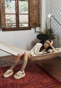 Beige hangmat die een persoon ondersteunt die een crèmekleurig shirt, tan schoenen met witte banden en beige gestructureerde sokken draagt, op een rood patroon tapijt.