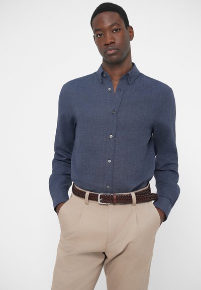 Jūras zilas pogas krekls ar teksturētu audumu, apvienots ar gaiši bēšiem biksēm un austu brūnu jostu. Redzamu rakstu vai akcentu nav.