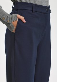 Tumši zilie bikses, izgatavotas no gludas auduma ar pielāgotu piegriezumu, ar sānu kabatām un pogu aizdari jostasvietā.