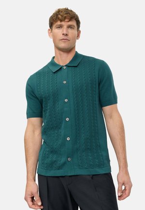 Mann trägt ein kurzärmliges, türkises Strickhemd mit Knöpfen, Kragen und strukturierten vertikalen Zopfmustern, kombiniert mit schwarzen Hosen.