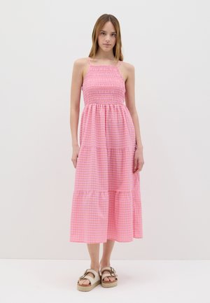 Vestito maxi rosa senza maniche con corpino arricciato, gonna a più strati e motivo a quadretti. Indossato con sandali leggeri, sfondo bianco semplice.