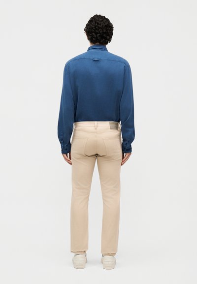 Άνδρας με μπλε μακρυμάνικο πουκάμισο, μπεζ παντελόνι και λευκά αθλητικά παπούτσια, στέκεται με την πλάτη προς το μέρος του θεατή, πάνω σε απλό λευκό φόντο.